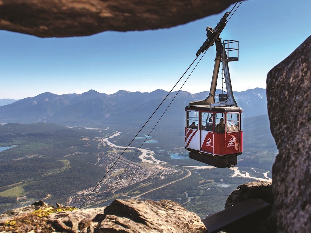 A red and white car is suspended on a cable high above the Jasper townsite, with mountains in the distance.