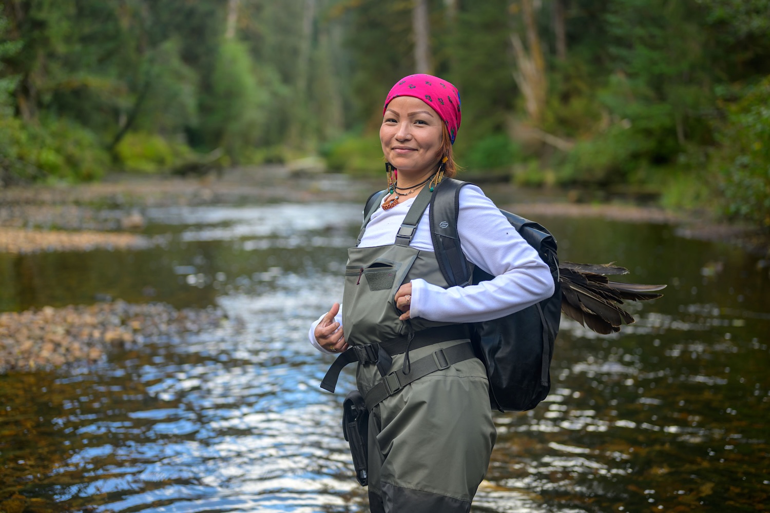 Santana Edgar has medium-light skin tone and is wearing a hot-pink bandana on her hair. She is wearing hip waders and standing in a river. 