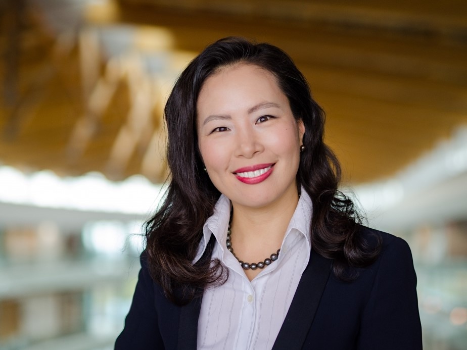 A woman with light skin tone and long black hair, wearing a black blazer over a white pinstriped button-up shirt, smiles at the camera.