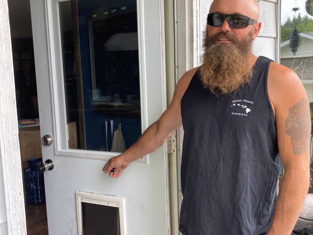 A light-skinned man with a shaved head and long beard points at a dog door. He wears a blue muscle shirt.
