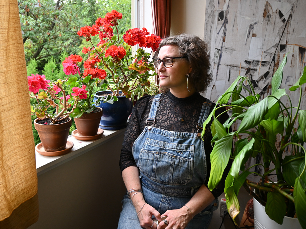 Riel Hahn sits beside her window near her red geranium flowers. She has light skin tone and curly, shoulder-length salt-and-pepper hair. She is wearing denim overalls and a black lace top.