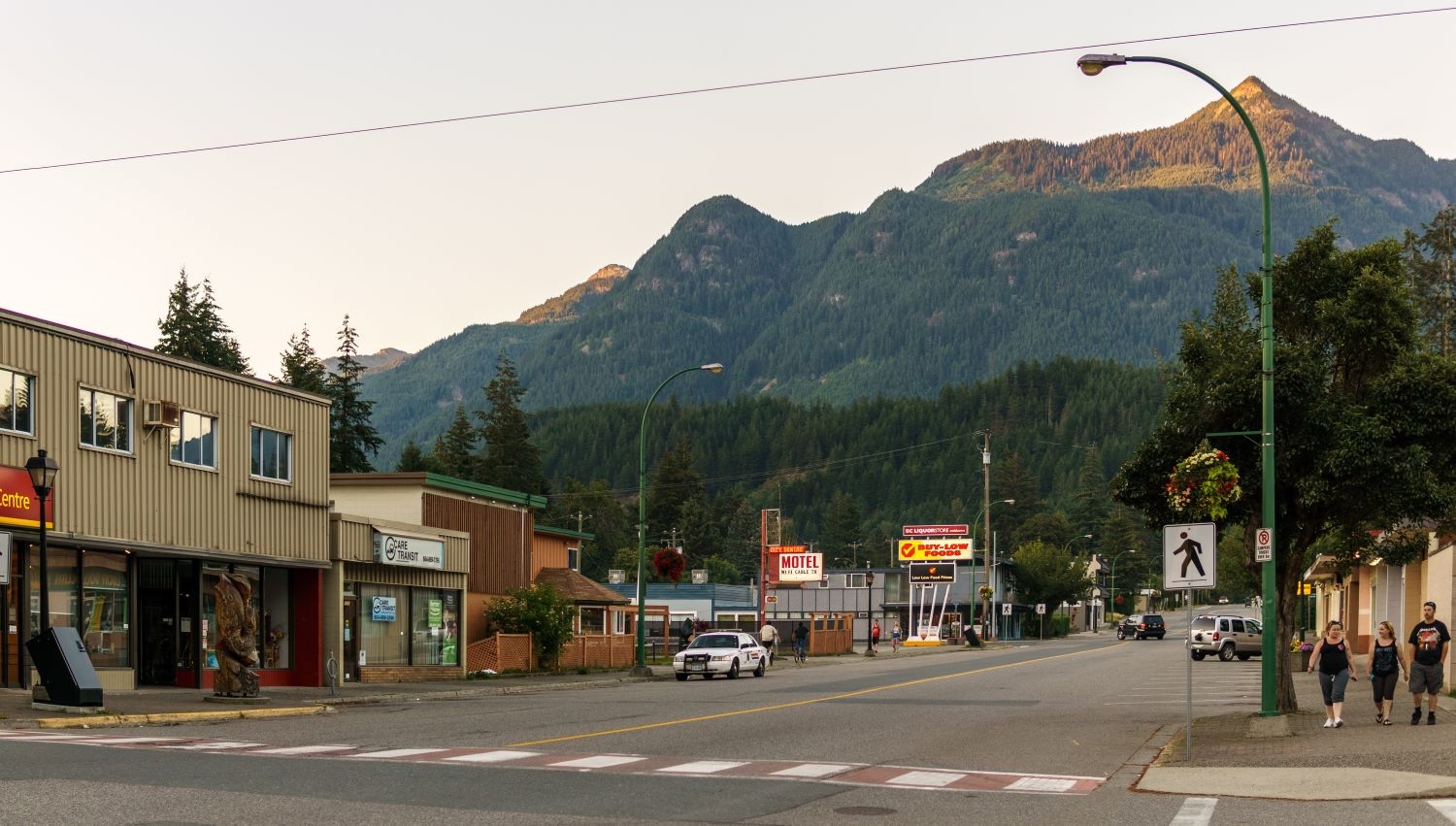 Three people stroll down a sidewalk in an otherwise deserted main drag of a small town with a few lit signs at dusk. In the background a bluish mountain rises.