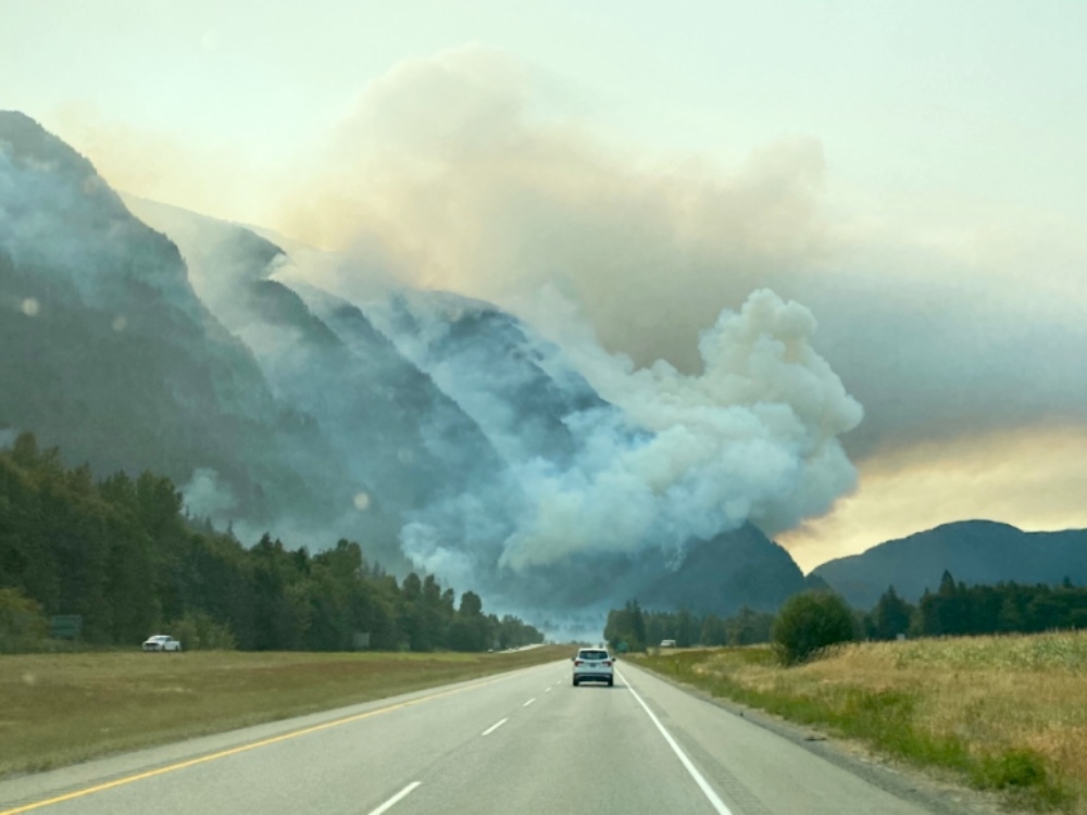 A photo taken from a highway showing smoke from a wildfire billowing from a mountainside ahead.