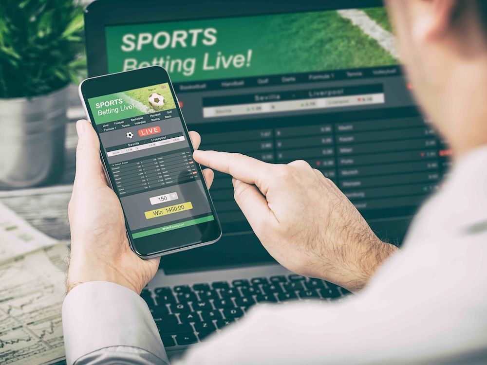 A person uses a phone with a sports gambling site displayed on the screen, in front of a laptop also open to a betting site.