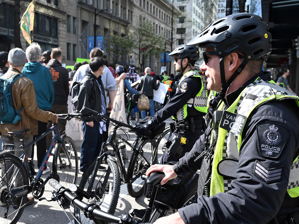 Two police officers wearing bike helmets and standing beside their bikes look on as people march to commemorate the nine-year anniversary of when B.C. declared a public health emergency to combat overdose fatalities.