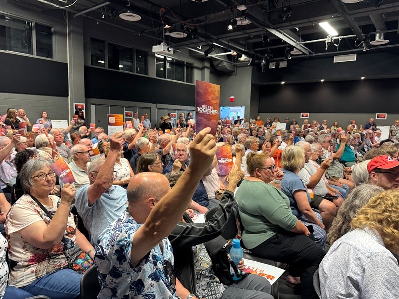 A packed auditorium of mostly older people. In the foreground someone holds up a pamphlet that says &lsquo;Better Together.&rsquo;