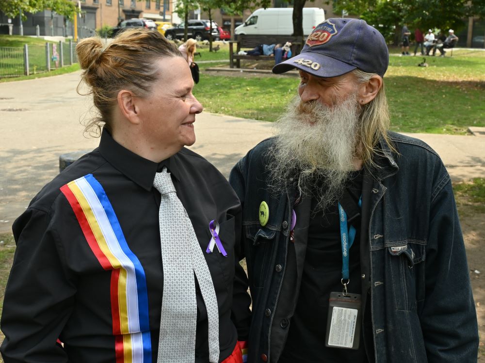 A white middle-aged woman with reddish hair wearing a black shirt and light grey tie and rainbow suspenders smiles and speaks with a senior man with long grey hair and a long grey beard wearing black clothes and a ball cap.