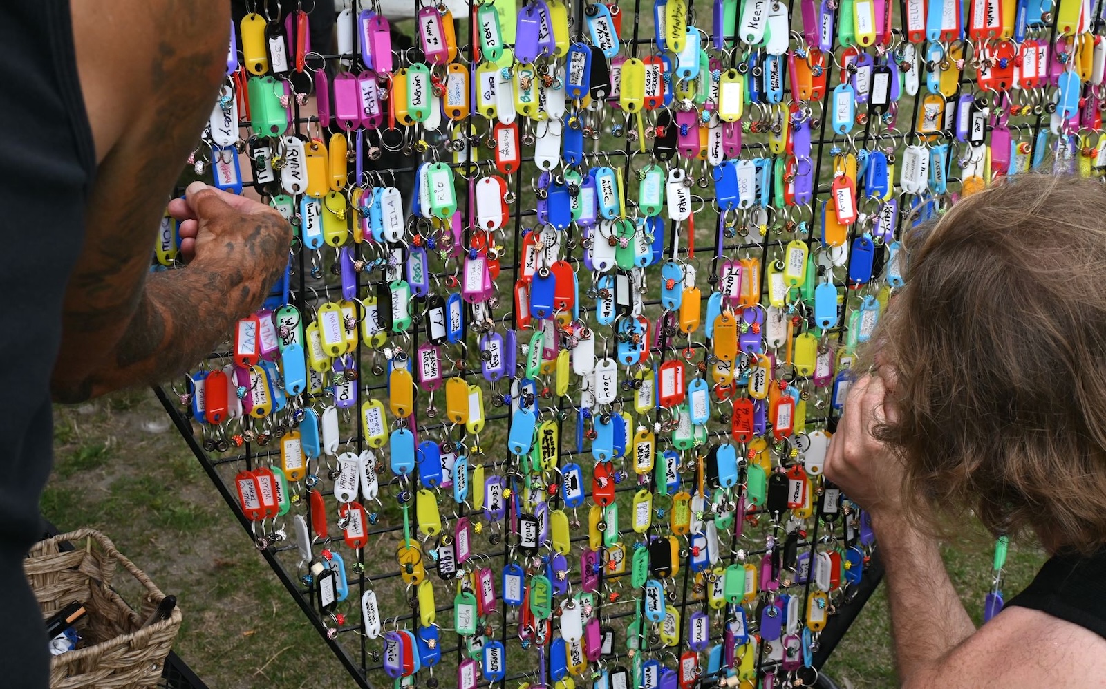 The partial backs of heads and arms and hands of two people are seen adding names on key chain-like tags to a colourful board displaying hundreds of other names.