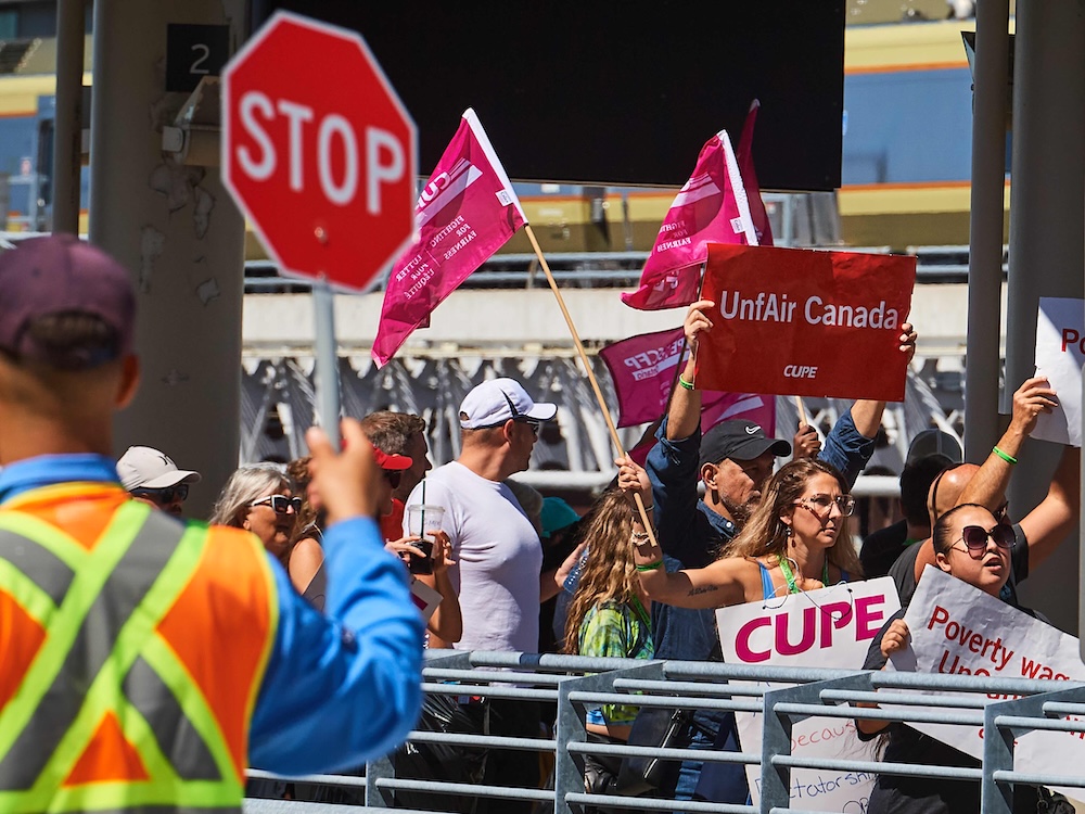 A person in the foreground holds up a Stop sign as people march in the background holding signs that say &lsquo;UnfAir Canada&rsquo; and &lsquo;CUPE&rsquo; outside an airport.