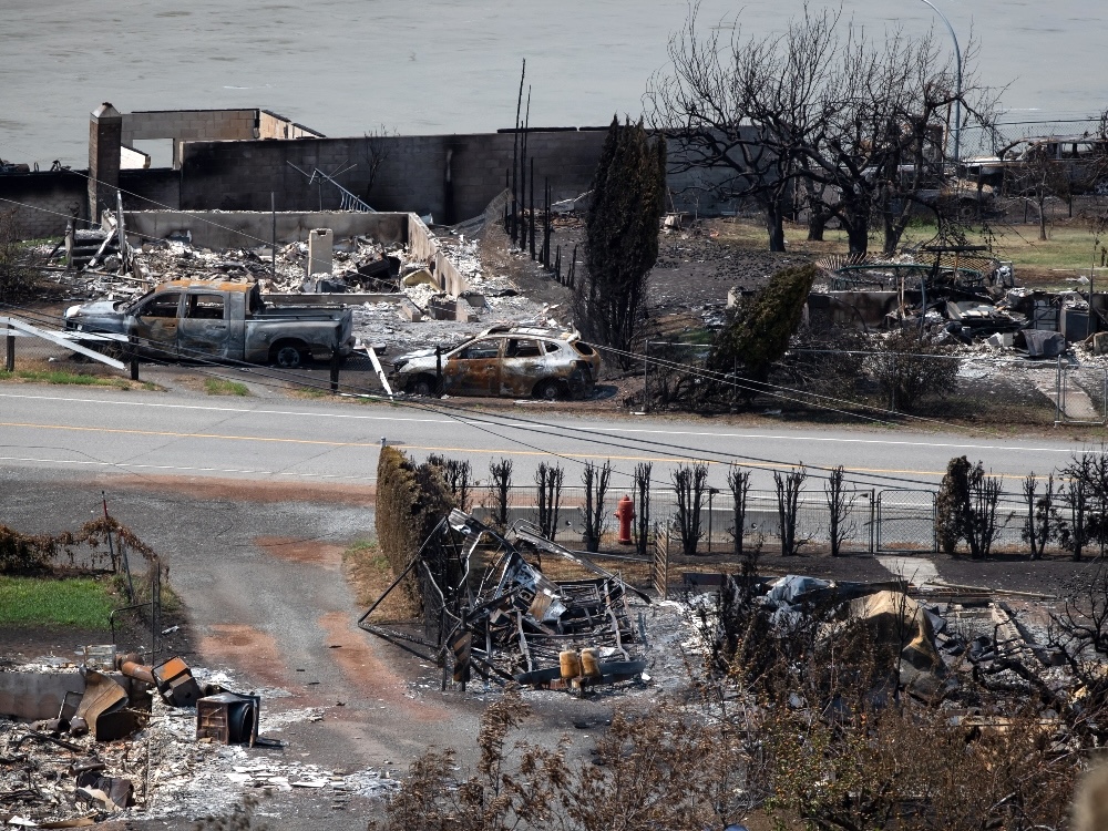 Burned-out properties line a road travelling through the small community of Lytton. The husks of burned vehicles are visible along the road. A river can be seen in the background.