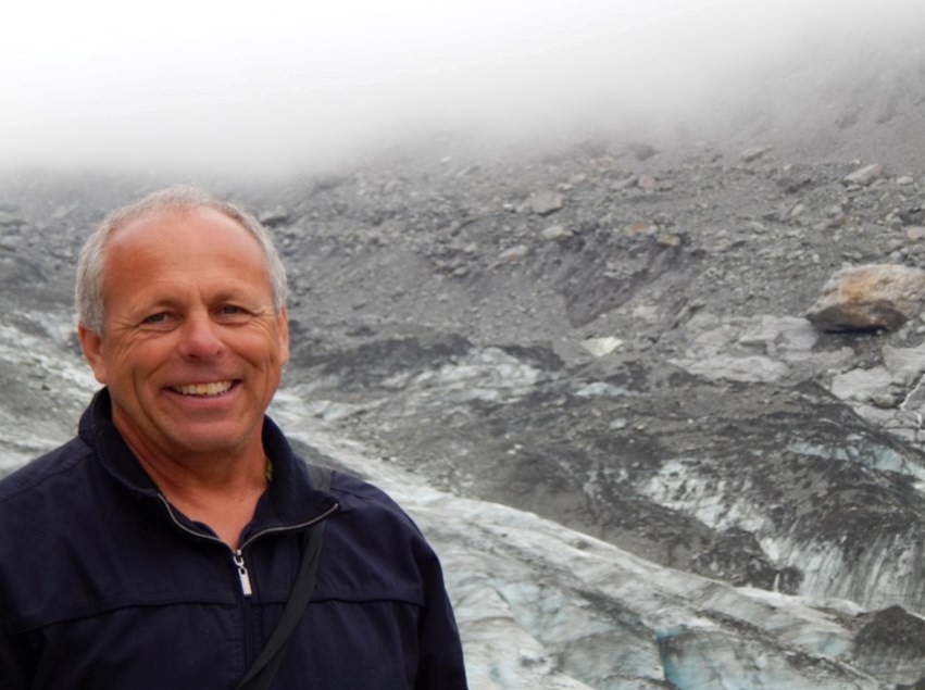 A smiling man with grey hair, wearing a blue jacket, stands near snow and rocks.