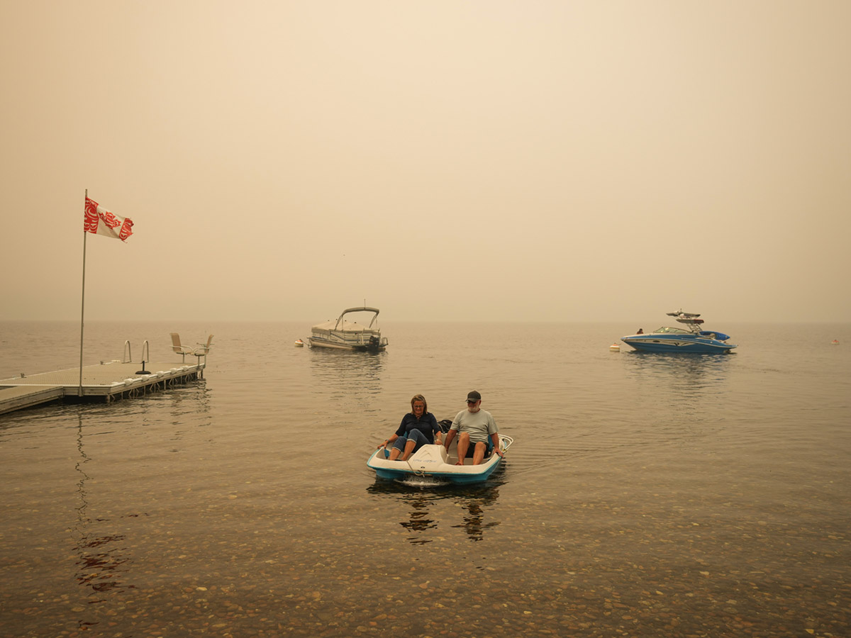 Two people sit in a paddleboat on a calm body of water. The air and water are tinted with the greasy orange-grey of wildfire smoke. Two boats sit with anchors down in the background. The smoke is so thick the far shore is not visible.