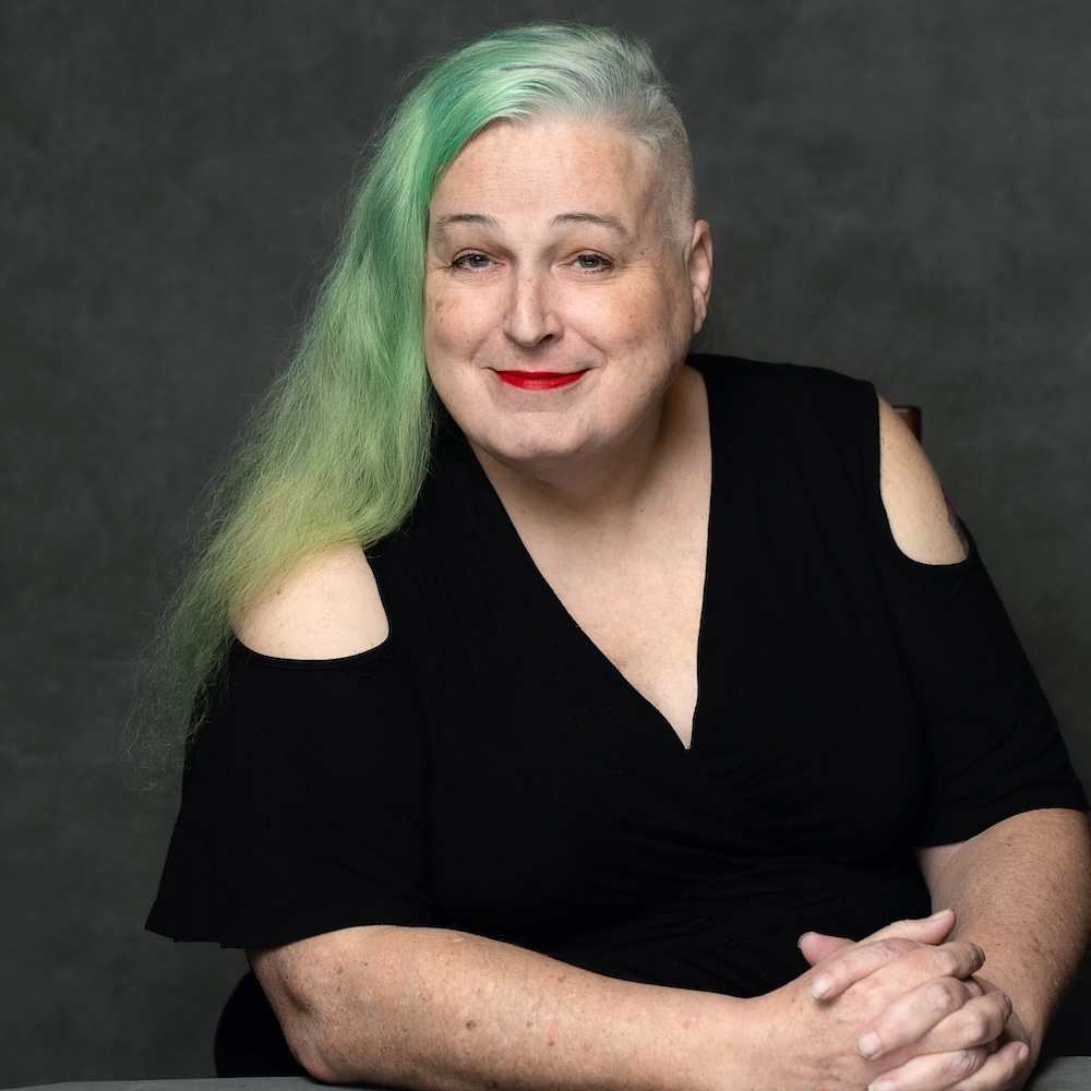 A headshot shows a woman with long green hair and light skin tone. She is wearing red lipstick and raising her eyebrows at the camera.