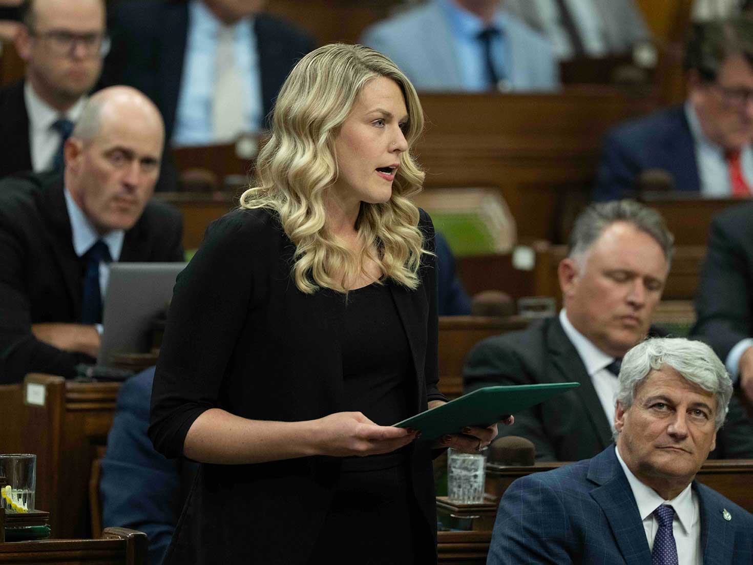 A light-skinned woman with blonde hair stands in at a desk in the House of Commons.