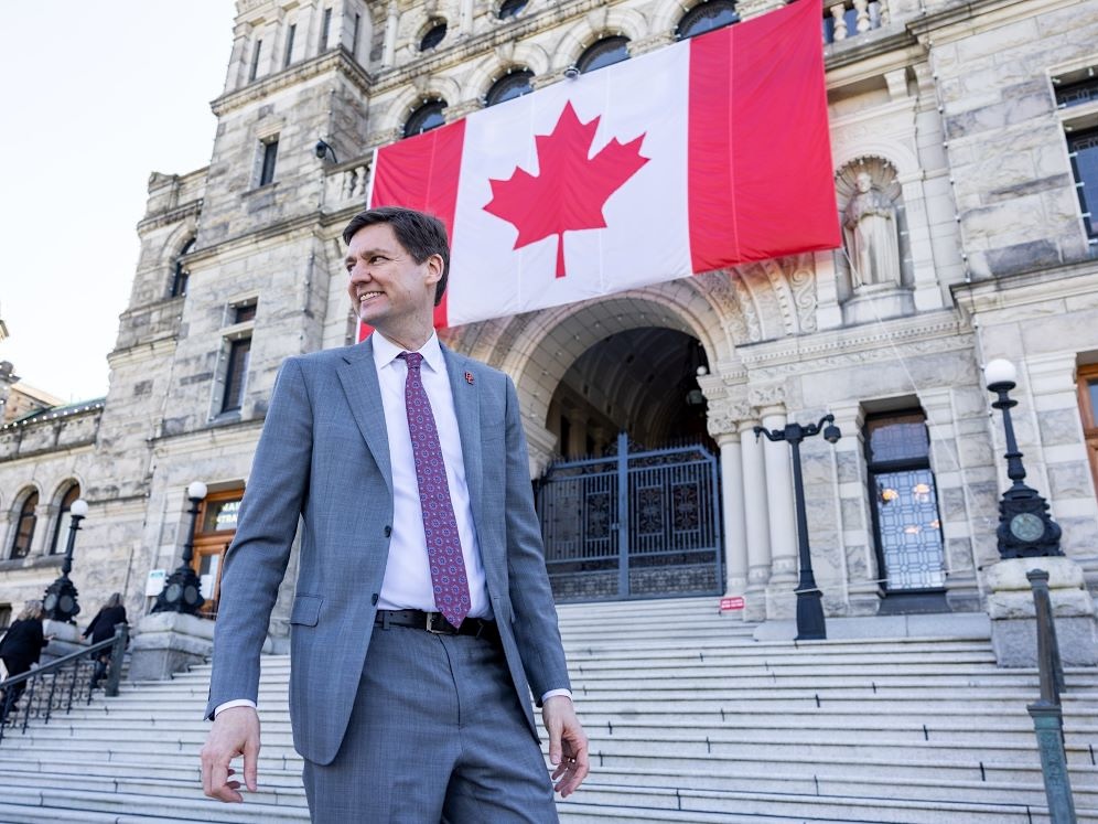 David Eby smiles in front of the legislature. Behind him a very large Canadian flag hangs on the building.