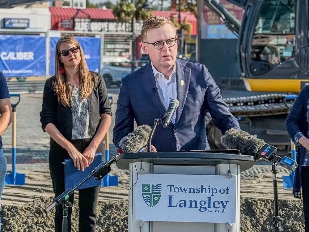 A light-skinned man with brown hair and glasses stands at a lectern that reads &lsquo;Township of Langley&rsquo; in front of construction equipment. Three other people can be seen standing behind him.