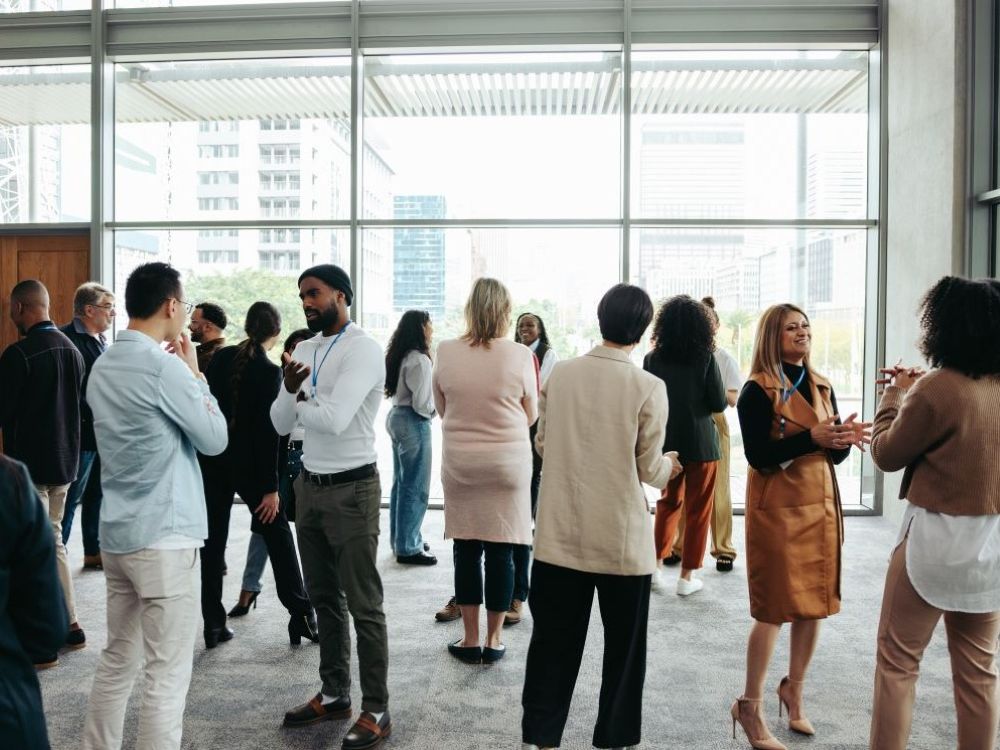 A sparse crowd of nicely dressed people of varying genders, ages and skin tones converse among each other in a well-lit room with grey carpet and floor-to-ceiling windows.