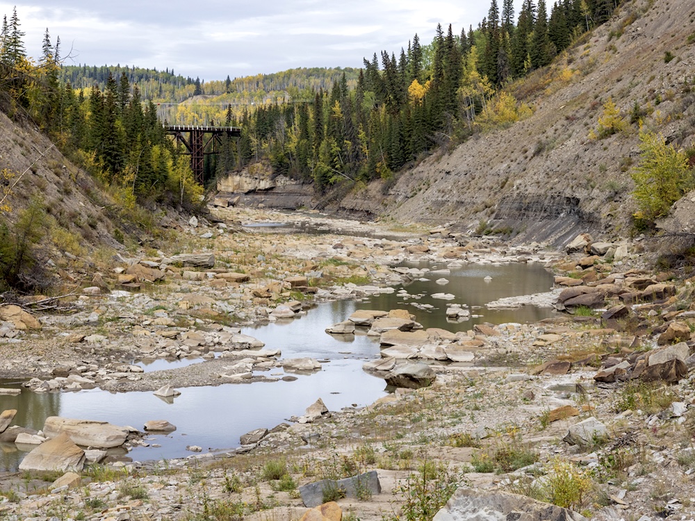 A river valley with an old trestle bridge in the background. The river&rsquo;s flow is very low, and many rocks are visible above the water line.