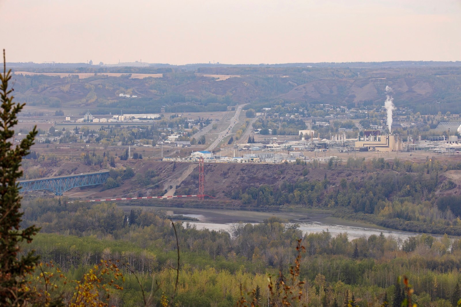 A view across the Peace River, which sits in a river valley. Across the valley is a large gas plant giving off a plume of smoke.