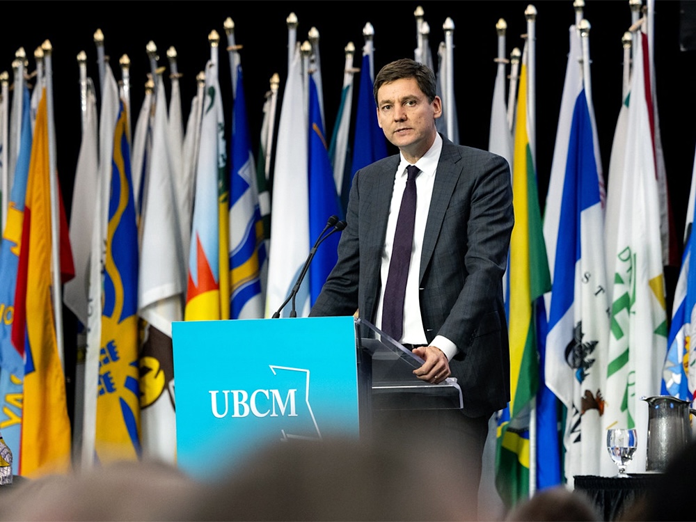 Premier David Eby stands on a stage with flags from municipalities across the province behind him.