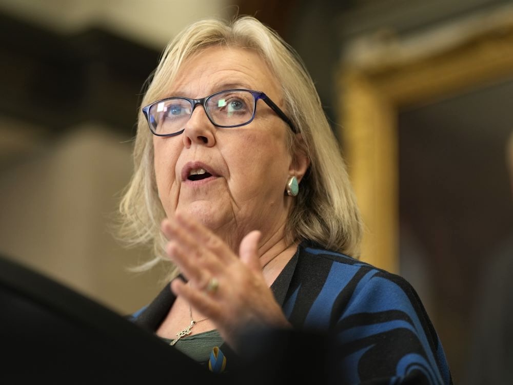 Elizabeth May speaks in the foyer of the House of Commons. She has light skin tone, a blond-grey bob and blue-black glasses.