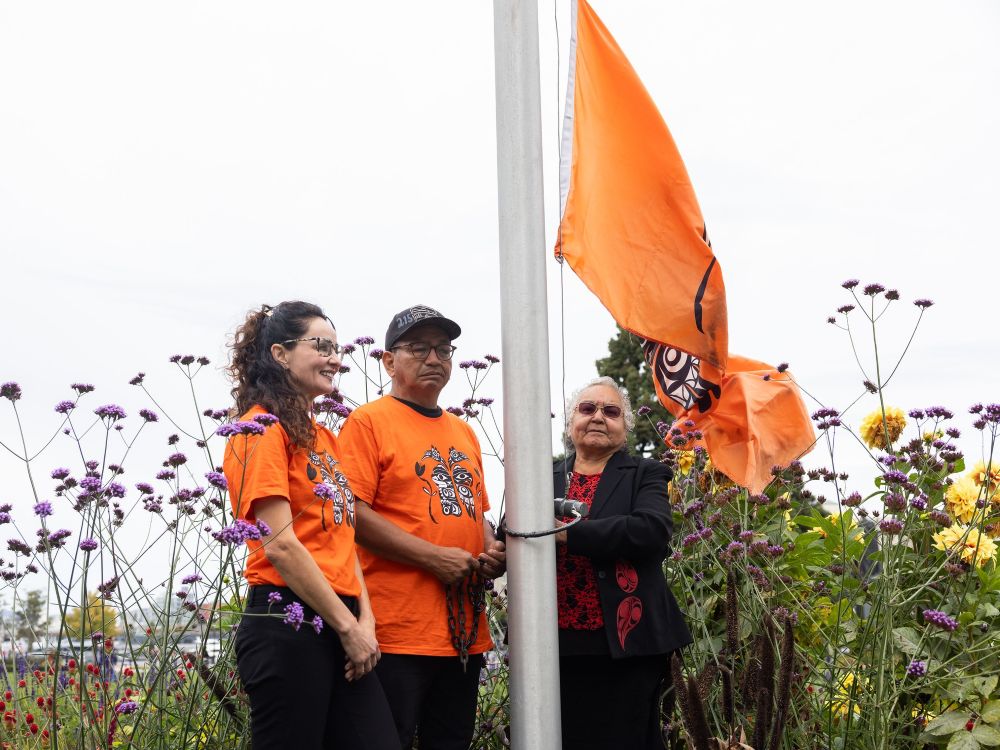 Two Indigenous people in orange shirts and another in a black jacket and red shirt raise an orange flag.
