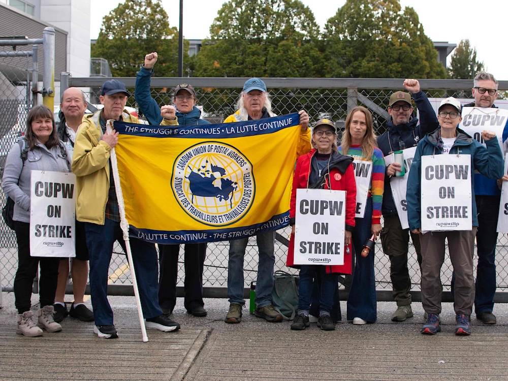 About a dozen people stand in front of a chain-link fence, holding signs with messages like &lsquo;CUPW on strike.&rsquo;