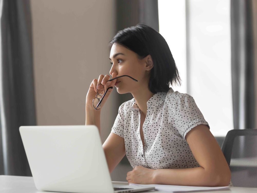 A woman with short dark hair sits at a desk with a laptop computer. She holds her glasses in her hands, looking to the middle distance. She is wearing a white patterned short-sleeved button-down shirt and there are large windows with grey drapes behind her on a cloudy day.