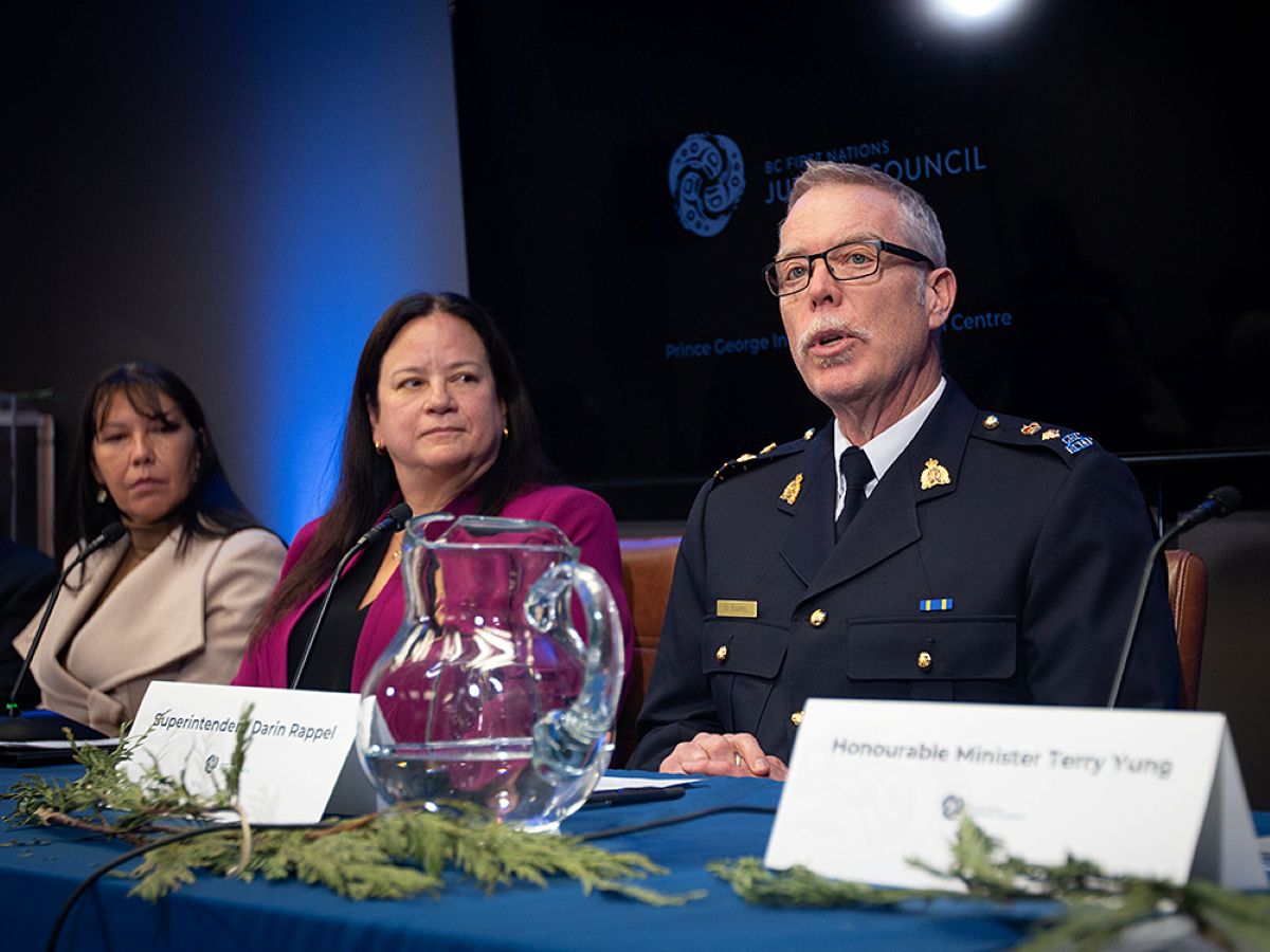 A man with short, grey hair and glasses sits at a table and speaks. He wears a dark uniform. Two women look on.