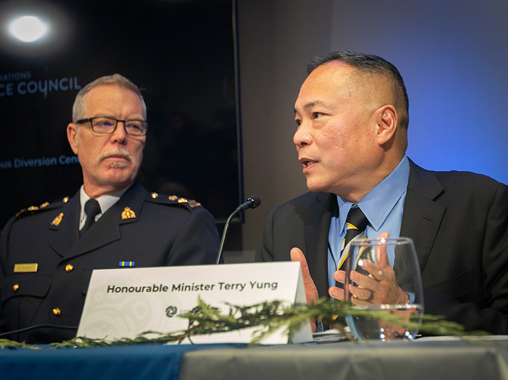 A man with short, dark hair sits at a table and speaks into a microphone. There is a water glass, a cedar bough and a name tag on the table that says &ldquo;Honourable Minister Terry Yung.&rdquo;