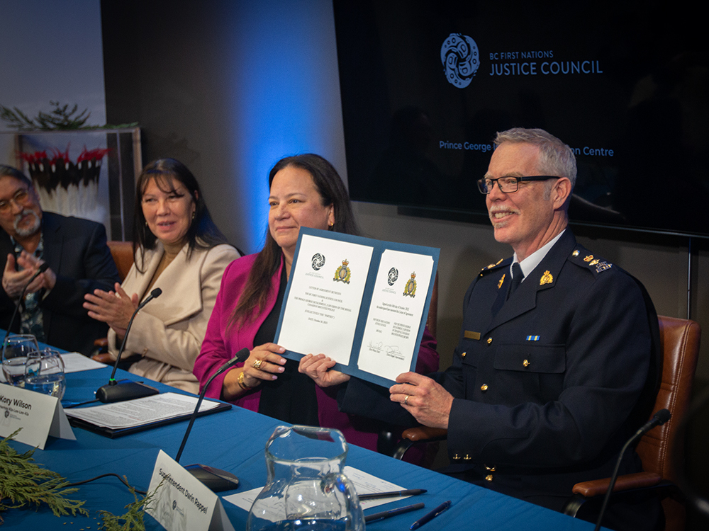 A woman with dark hair and a pink jacket sits at a table next to a man with short, grey hair and glasses, and wearing a dark uniform. They hold up a signed document.