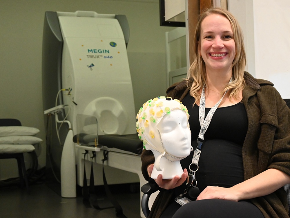 A woman with light skin tone and shoulder-length blond hair holds a Styrofoam dummy head with sensors attached. Behind her is the MEG, a big white and grey machine with a head-shaped indentation.