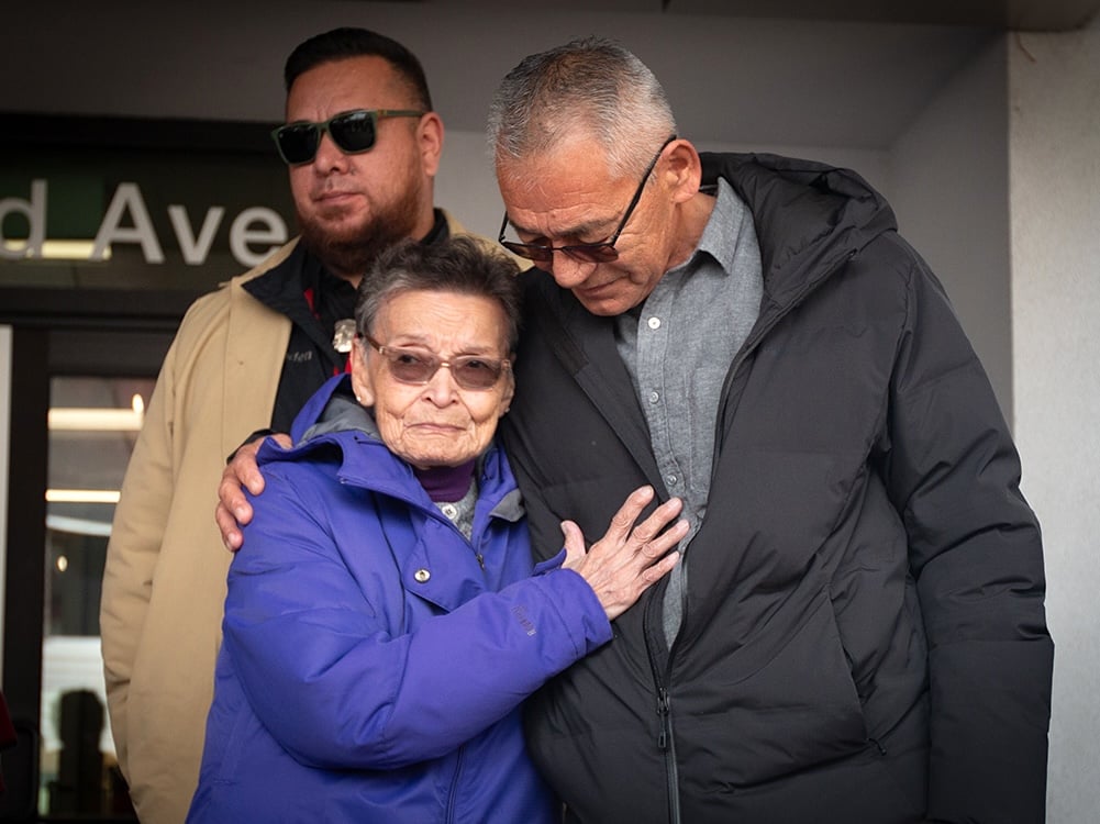 An elderly woman in a purple coat and glasses places a hand on the chest of a taller man in a black jacket and glasses, who puts an arm around her shoulder.