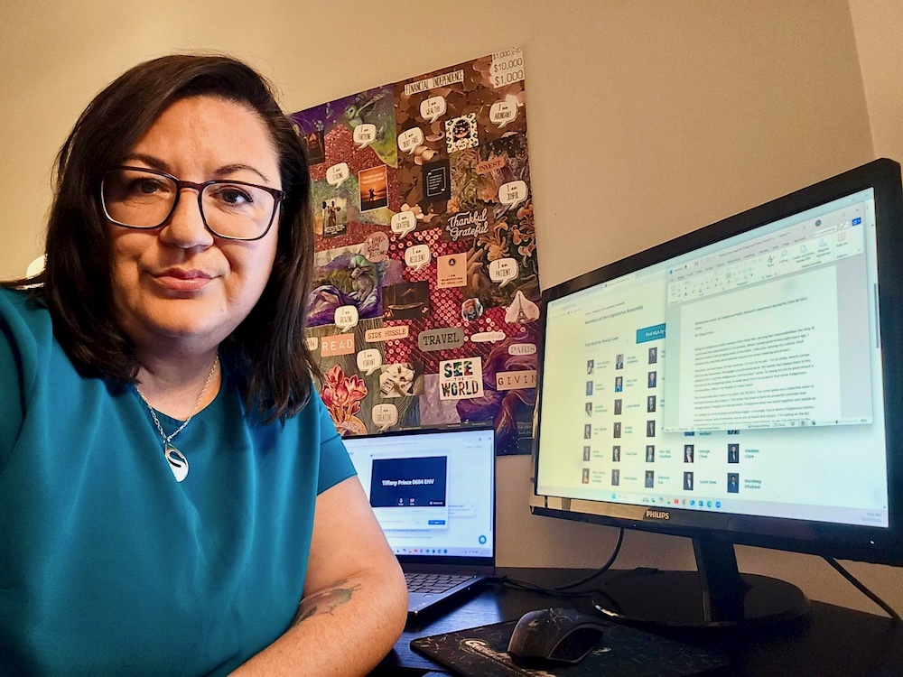 A woman with long brown hair and glasses looks at the camera. She sits in front of a desktop computer and a laptop.