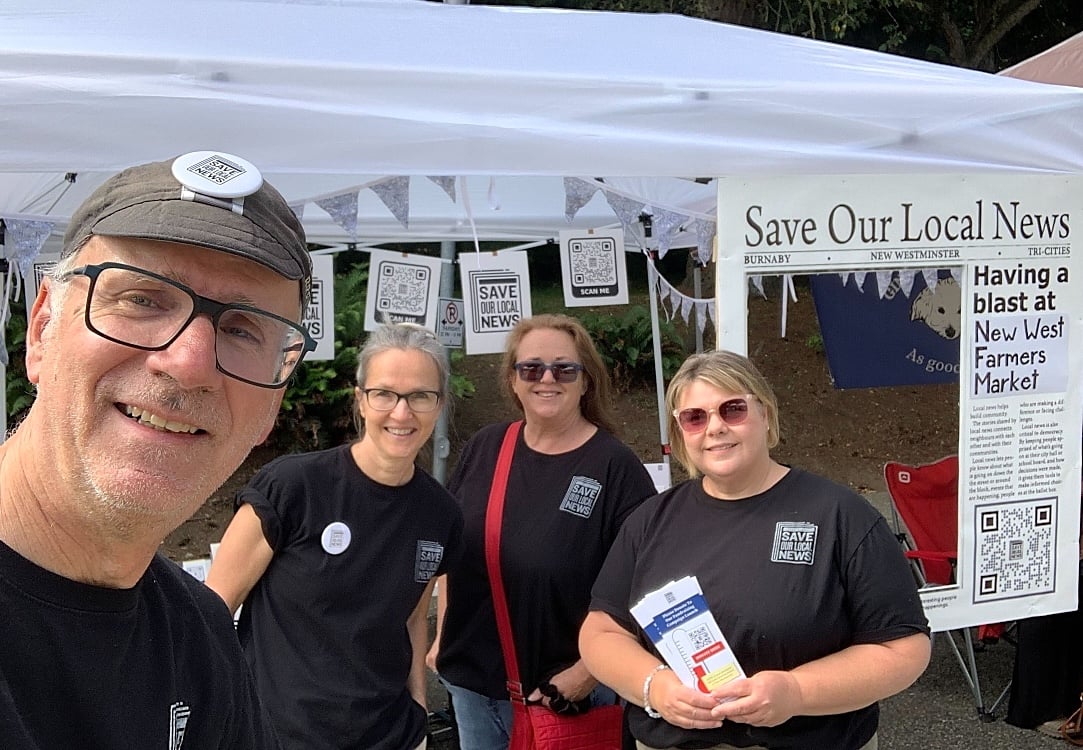 A selfie with Mario Bartel, a fair-skinned man wearing a hat, in the foreground and his three co-workers behind him next to a ‘Save Our Local News’ tent and display.