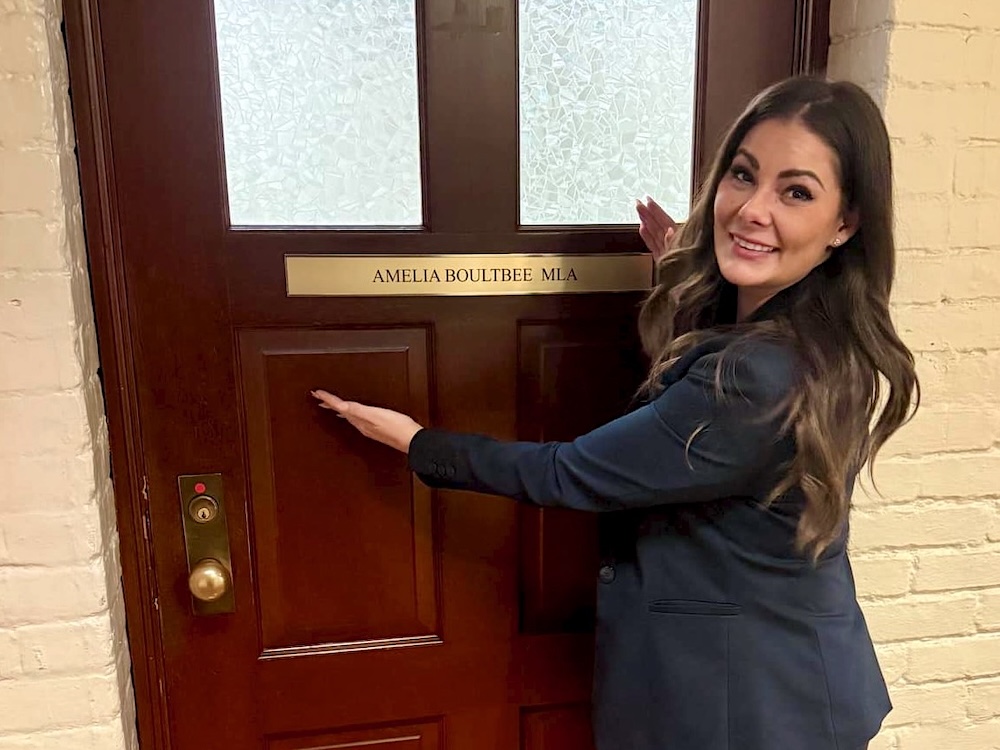 A young woman with long, wavy brown hair wearing a navy blue pantsuit stands in front of an office door in the basement of the B.C. legislature.