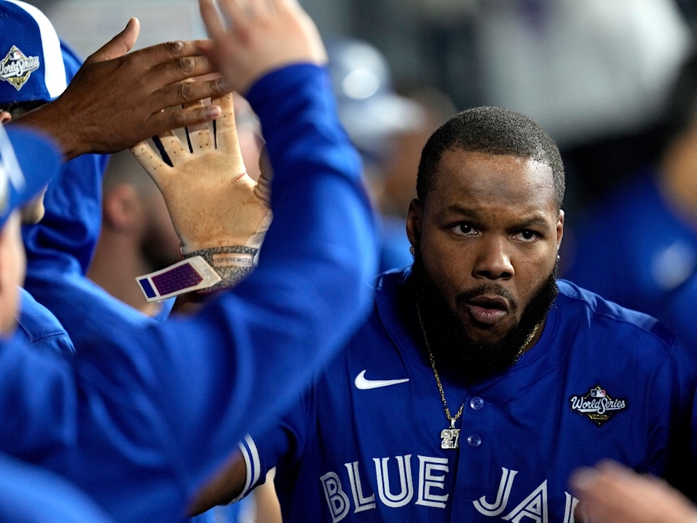 Vladimir Guerrero Jr. has dark skin, a prominent beard and close-cropped hair and is wearing a gold necklace with his number, 27. He is high-fiving teammates and has an intense look.