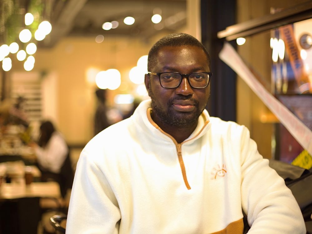 A man with dark skin tone wearing glasses and a white sweater sits in a coffee shop.