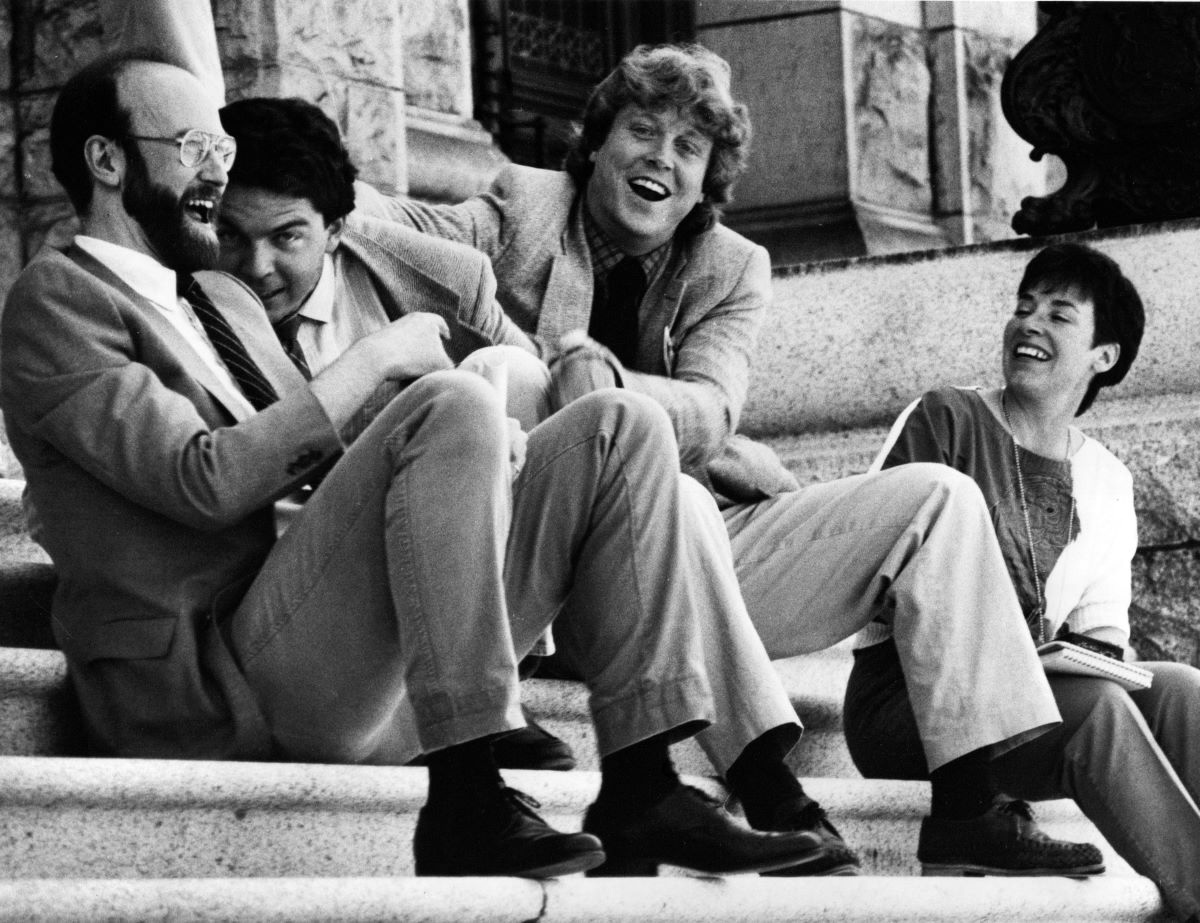 A black and white photo shows four journalists sitting and laughing on the steps of the B.C. legislature.