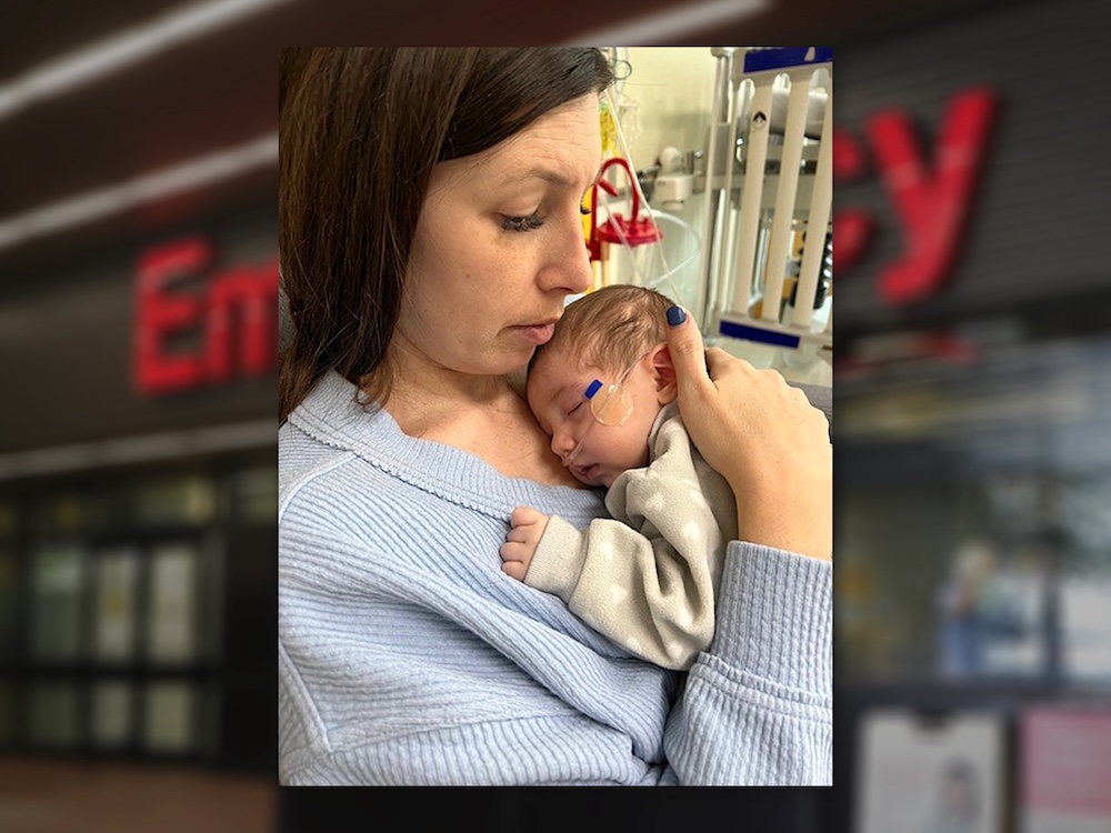 A woman with brown hair and a pale blue sweater holds an infant to her chest. The infant is wearing a cosy beige fleece top with white hearts on it and has an oxygen tube taped under her nose. Medical equipment can be seen in the background.