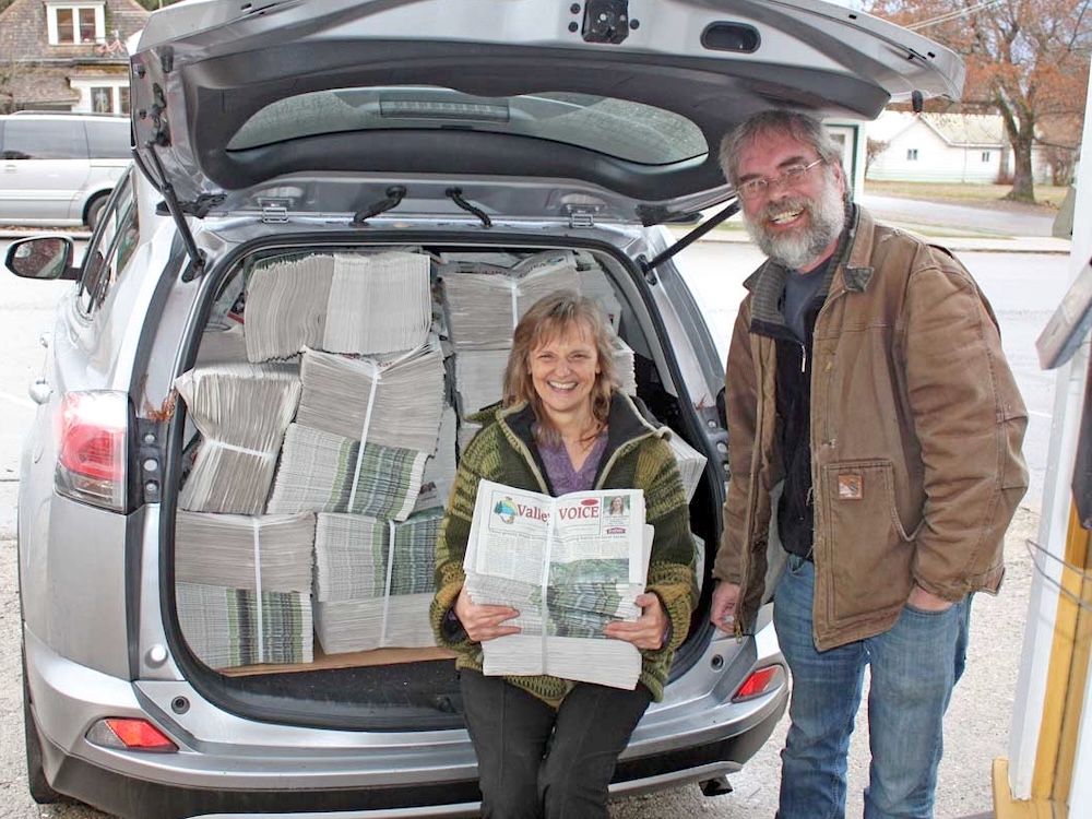 A smiling middle-aged couple stand in front of a van stuffed with newspapers ready for delivery. They&rsquo;re holding a copy of the Valley Voice.