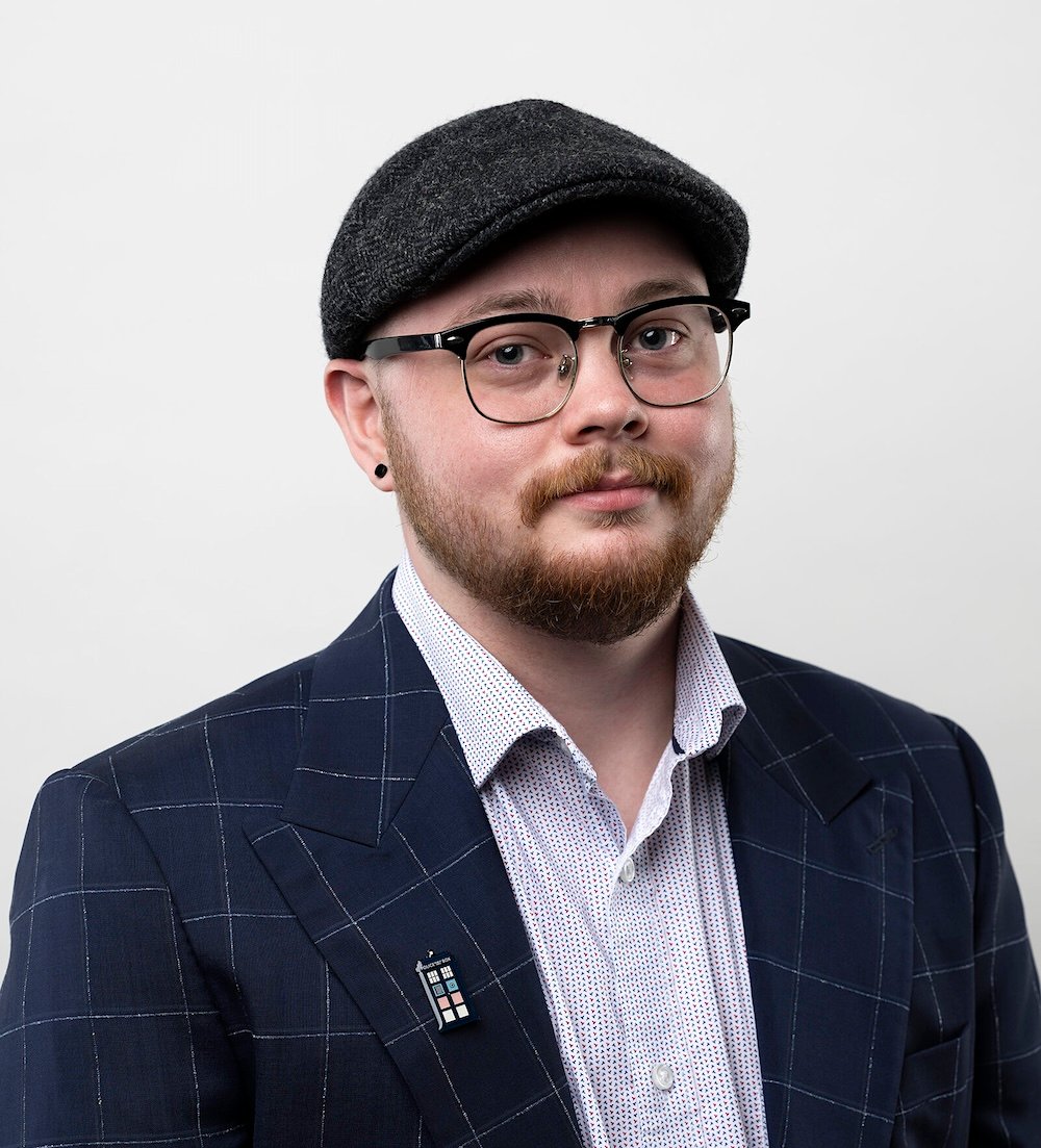 A man with light skin tone and an orange beard wears a plaid cap and looks at the camera.