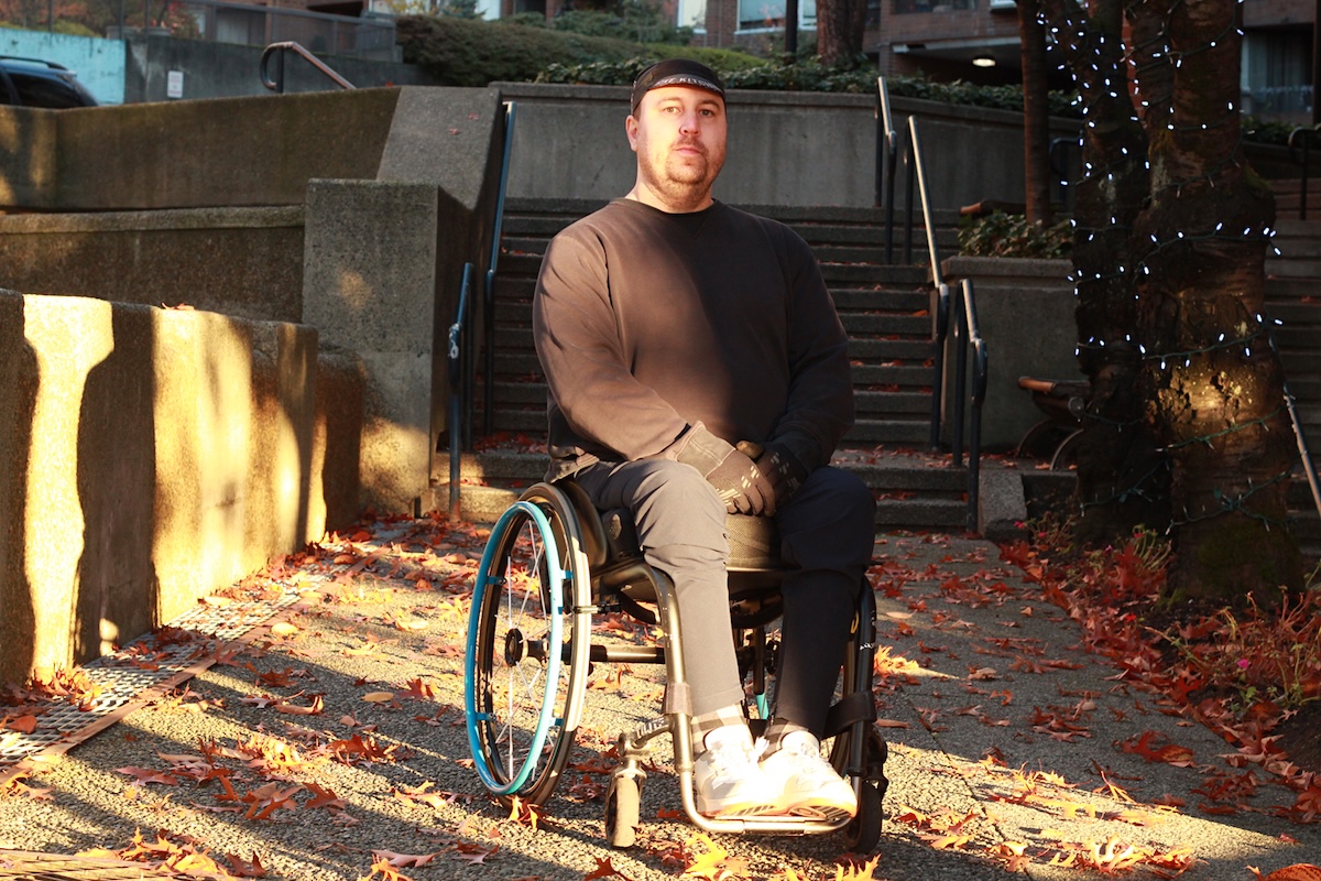 A person with light skin tone and short hair under a Bike Kitchen cycling cap wears all black. They are seated in a wheelchair.