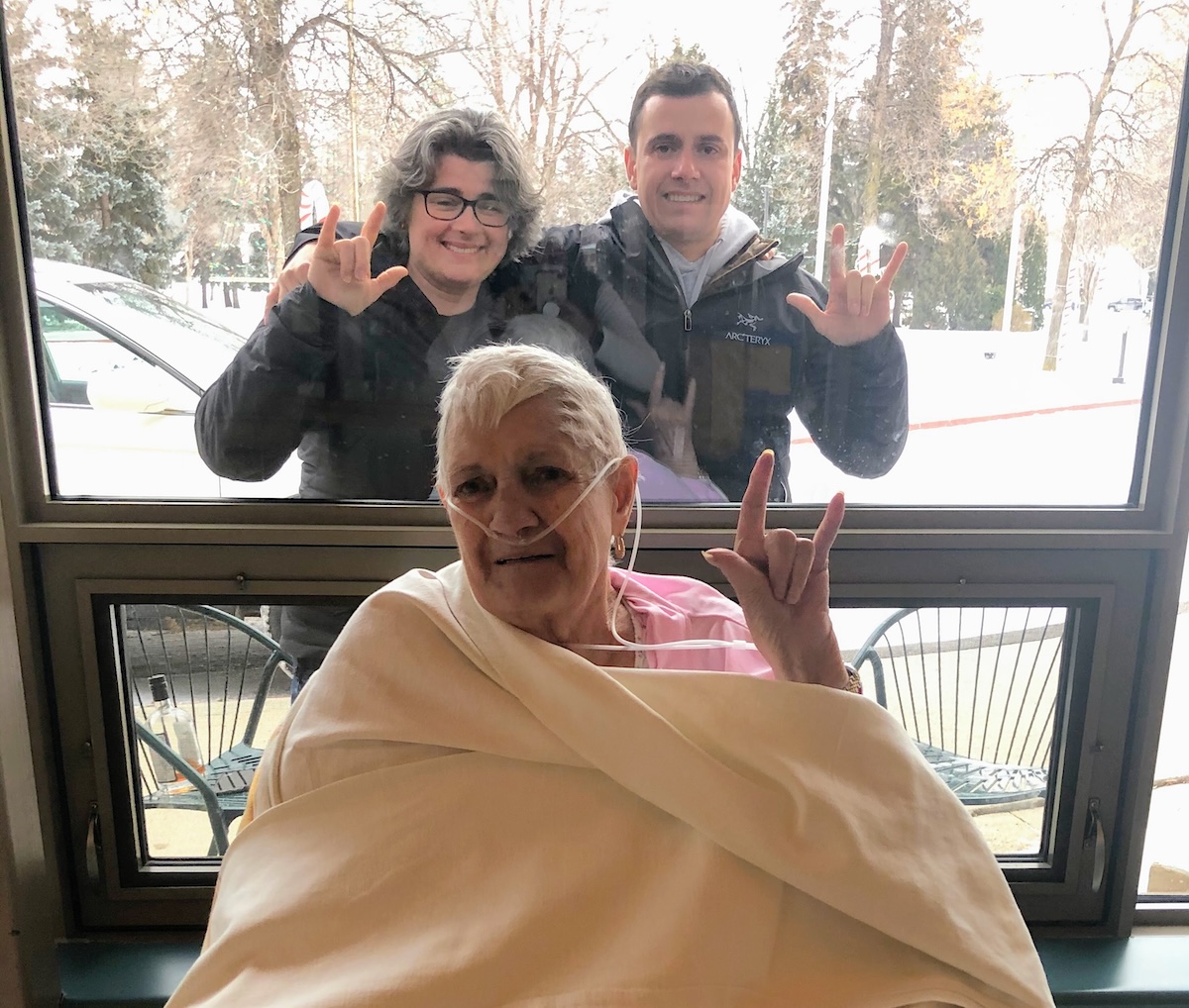 A woman and a man stand outside a window, both signing &lsquo;I love you.&rsquo; Inside, an older woman with grey hair, swaddled in blankets and using oxygen, makes the same sign.