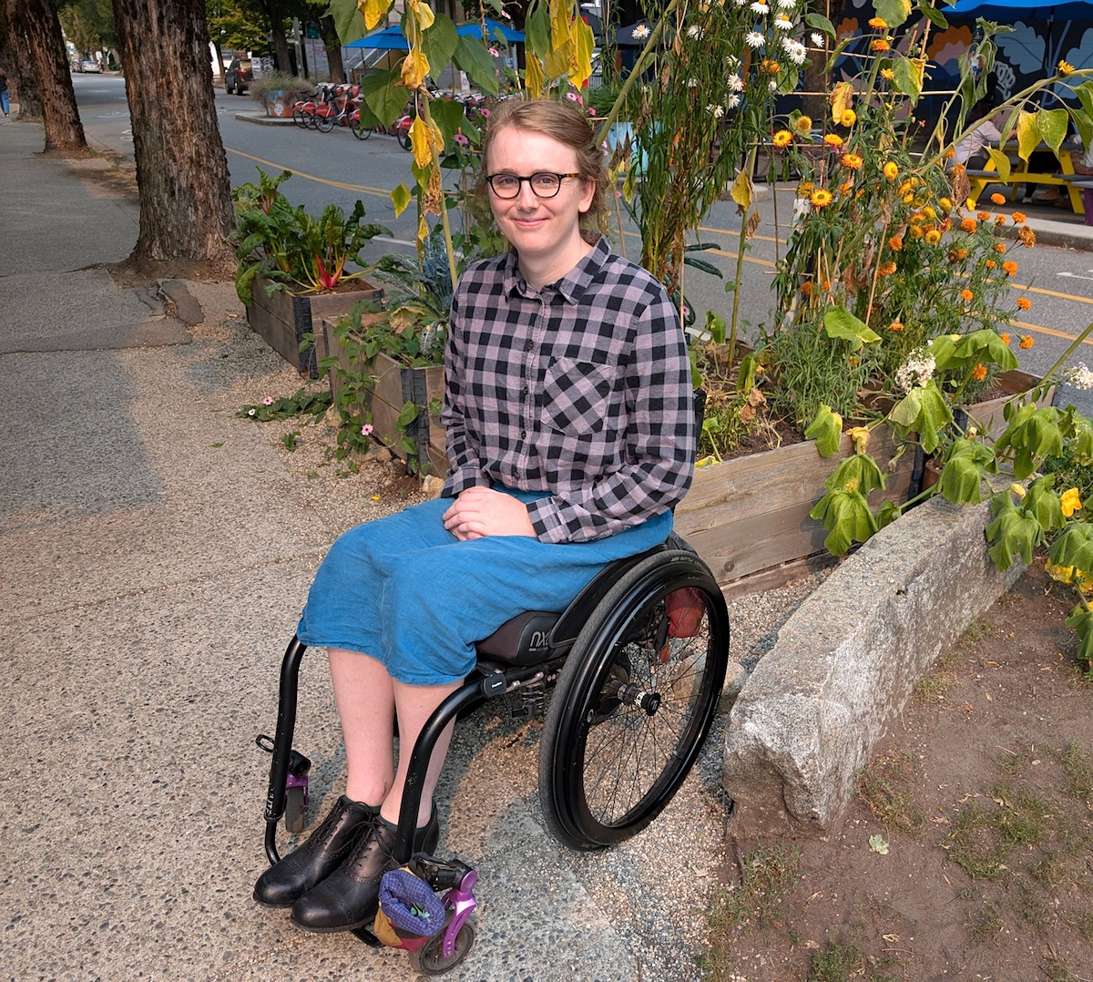 A woman with light skin and pulled-back blond hair smiles at the camera while posing in front of a late-summer garden. She&rsquo;s wearing black glasses, a purple and grey flannel shirt and a blue skirt. She&rsquo;s sitting in a manual wheelchair with her hands resting in her lap.