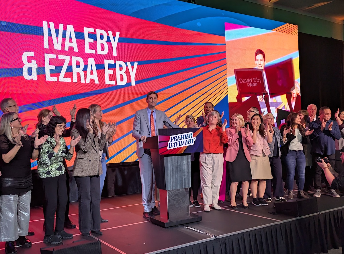 David Eby stands at a lectern on a stage, flanked by supporters, with a brightly lit colourful wall behind him displaying the words &lsquo;Iva Eby & Ezra Eby.&rsquo;