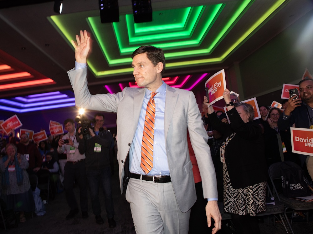 David Eby, a light-skinned man with short dark hair, waves to a crowd in front of coloured lights.