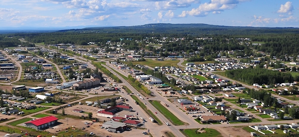 An aerial view of a small prairie town. A two-lane divided highway cuts through town, with shops and single-family homes on either side.