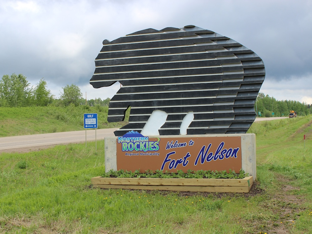 A sign marking Fort Nelson, shaped like a bear with horizontal stripes and surrounded by grass.