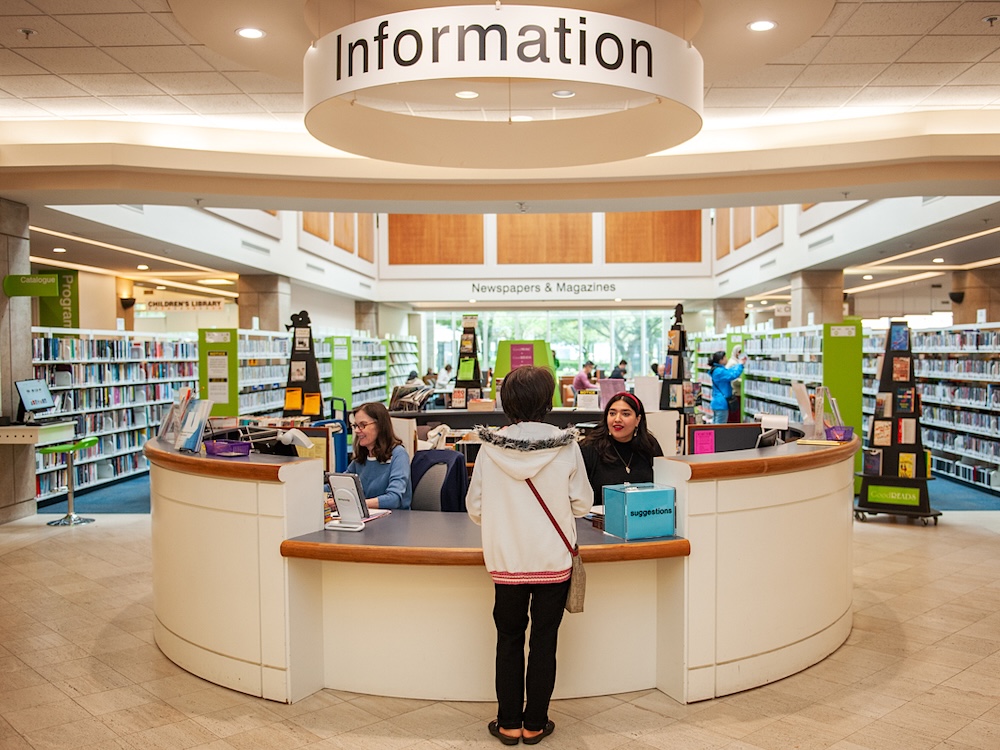 The interior of a Burnaby library branch, with two staff at a desk with a sign saying &lsquo;Information&rsquo; and shelves in the background.